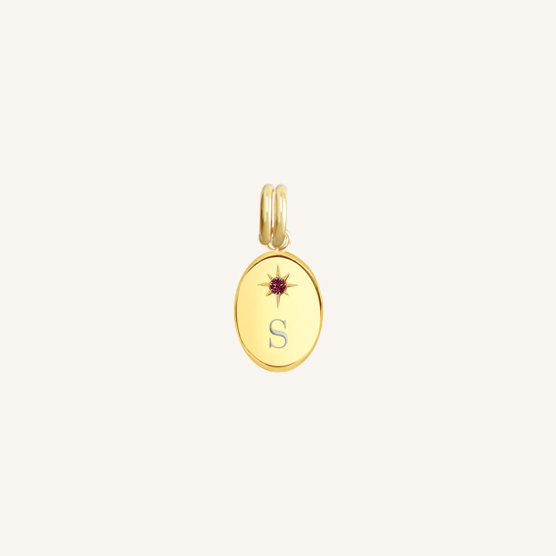 Gold pendant with a letter 'S' and red gemstone on a light beige background