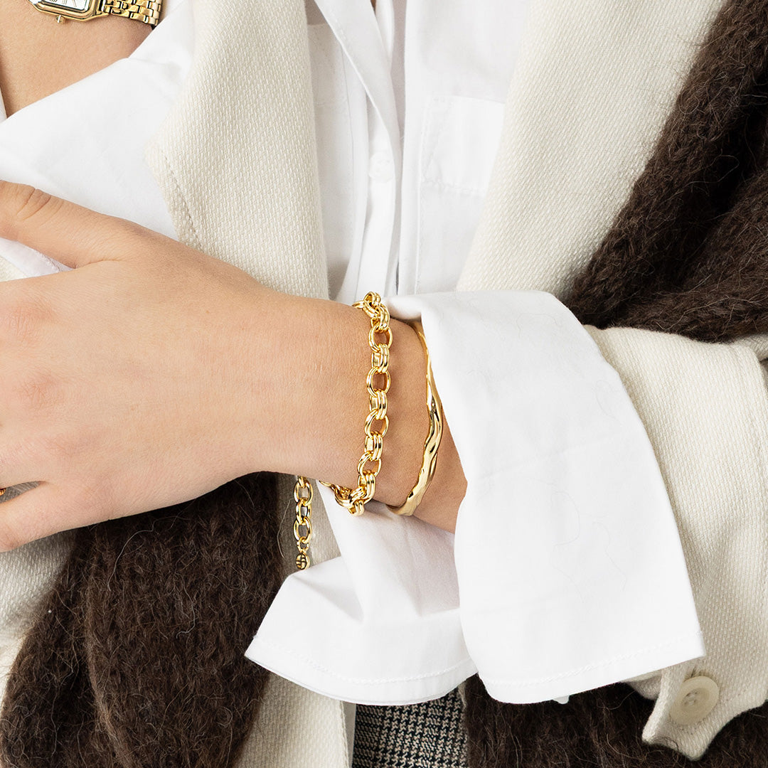 Gold chain bracelet on a wrist with a white shirt and brown sweater in the background