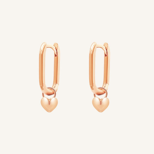 A pair of rose-tone hoop earrings with heart charms attached to them, displayed against a white background.
