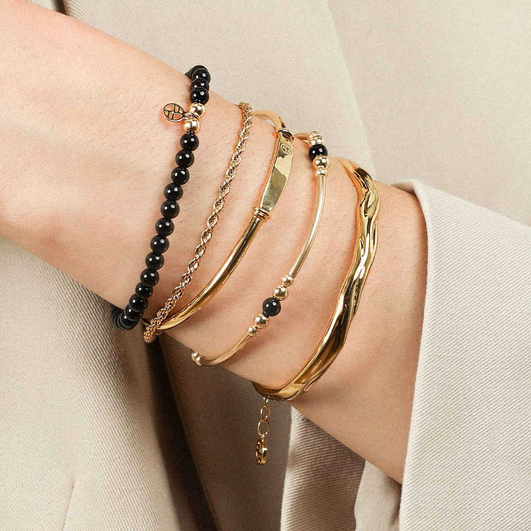 A black beaded bracelet made of black agate stones with a gold charm, worn on models wrist, stacked with other bracelets
