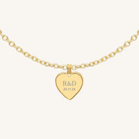 A gold heart-shaped necklace with a chain, featuring an engraved monogram initials and a date on the front.